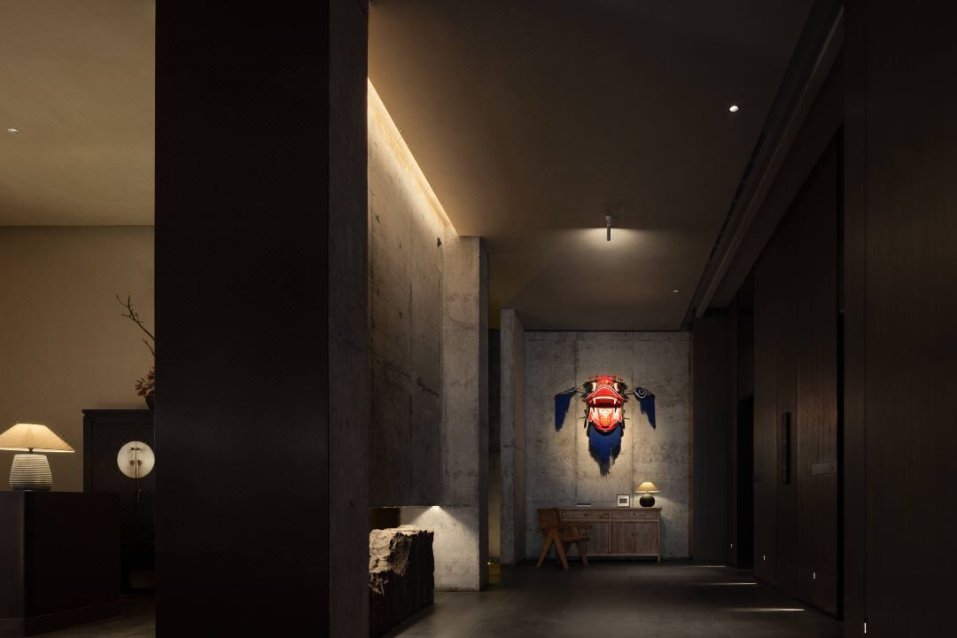 Un couloir faiblement éclairé, avec un mur en béton, un bureau sur lequel se trouve une lampe, et une œuvre d’art accrochée au mur.