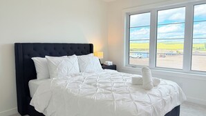 Ein ordentlich bezogenes Bett mit weißer Bettwäsche und einem dunklen Kopfteil in einem Zimmer mit einem großen Fenster.
