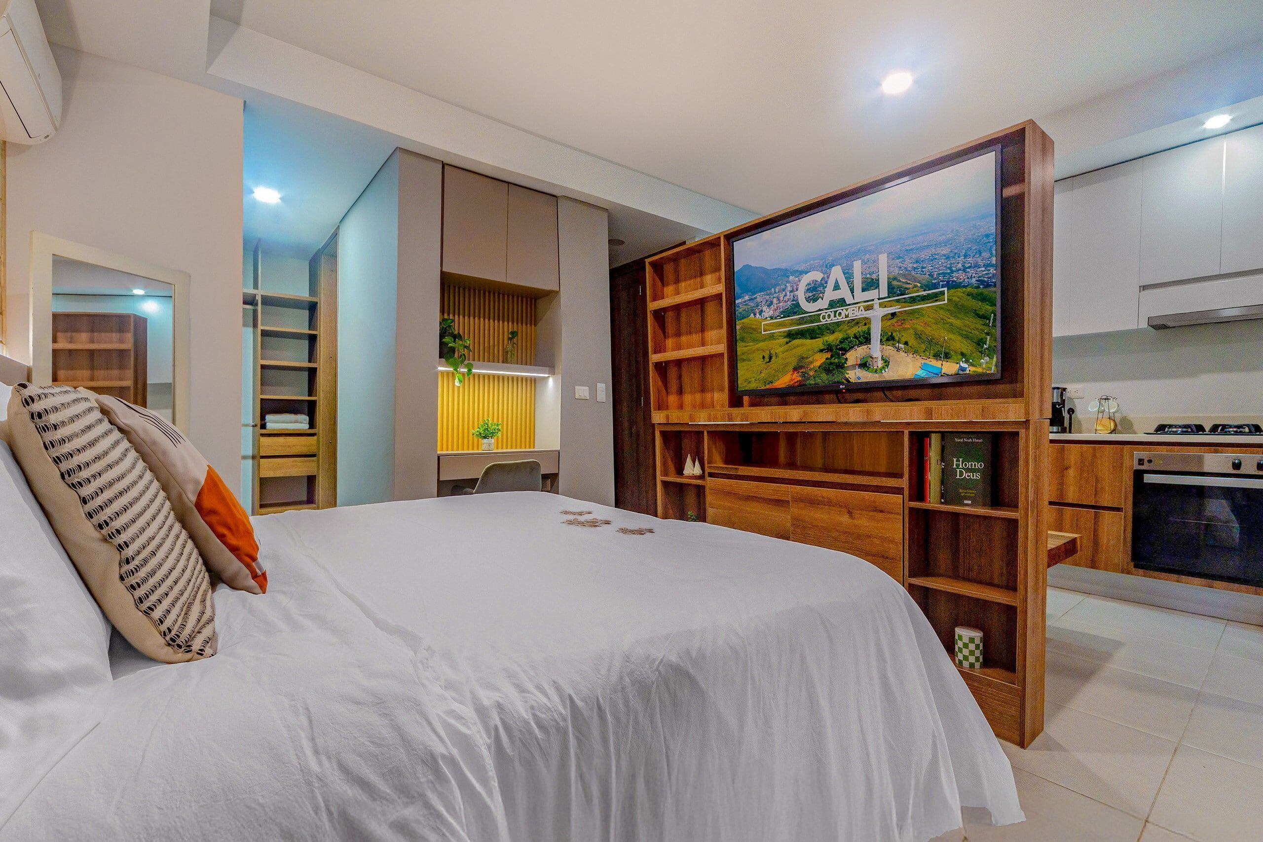 A bedroom with a bed, a TV mounted on a wooden shelf, and a kitchen area with a stove.