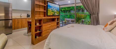 Ein modernes Schlafzimmer mit einem großen Bett, einer Holz-Unterhaltungseinheit mit Fernseher, einer Küchenzeile und einem Balkon mit Blick auf Grünflächen.