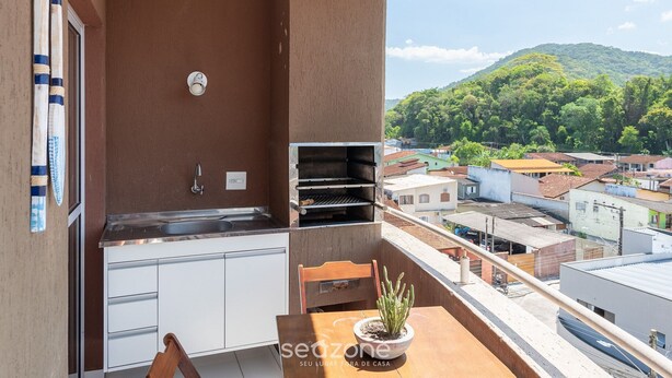 A balcony with a built-in grill, a small table with a potted plant, and a view of a cityscape and hills. - Apt w/ BBQ Near the Waterfront Ubatuba-sp Cdv042 (Ubatuba)