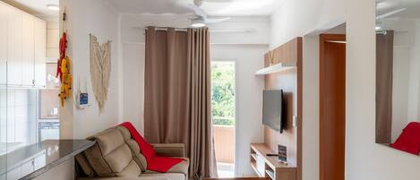 A modern living room with a sofa, a dining table, and a TV. There is a kitchen area with cabinets and a window.