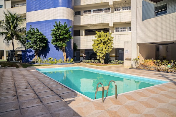 Una piscina con borde de baldosas, rodeada de edificios con balcones y árboles.