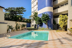 Una piscina con borde de baldosas, rodeada de vegetación y un edificio de fachada azul y blanca.