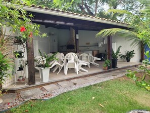 A covered outdoor seating area with white plastic chairs, potted plants, and a brick oven.