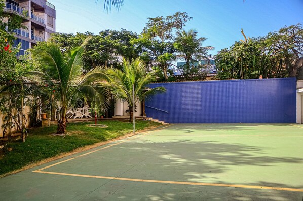 An outdoor tennis court with a blue wall, surrounded by greenery and a building in the background.