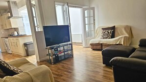 A living room with a sofa, a television on a stand, a kitchen area, and a door leading outside.