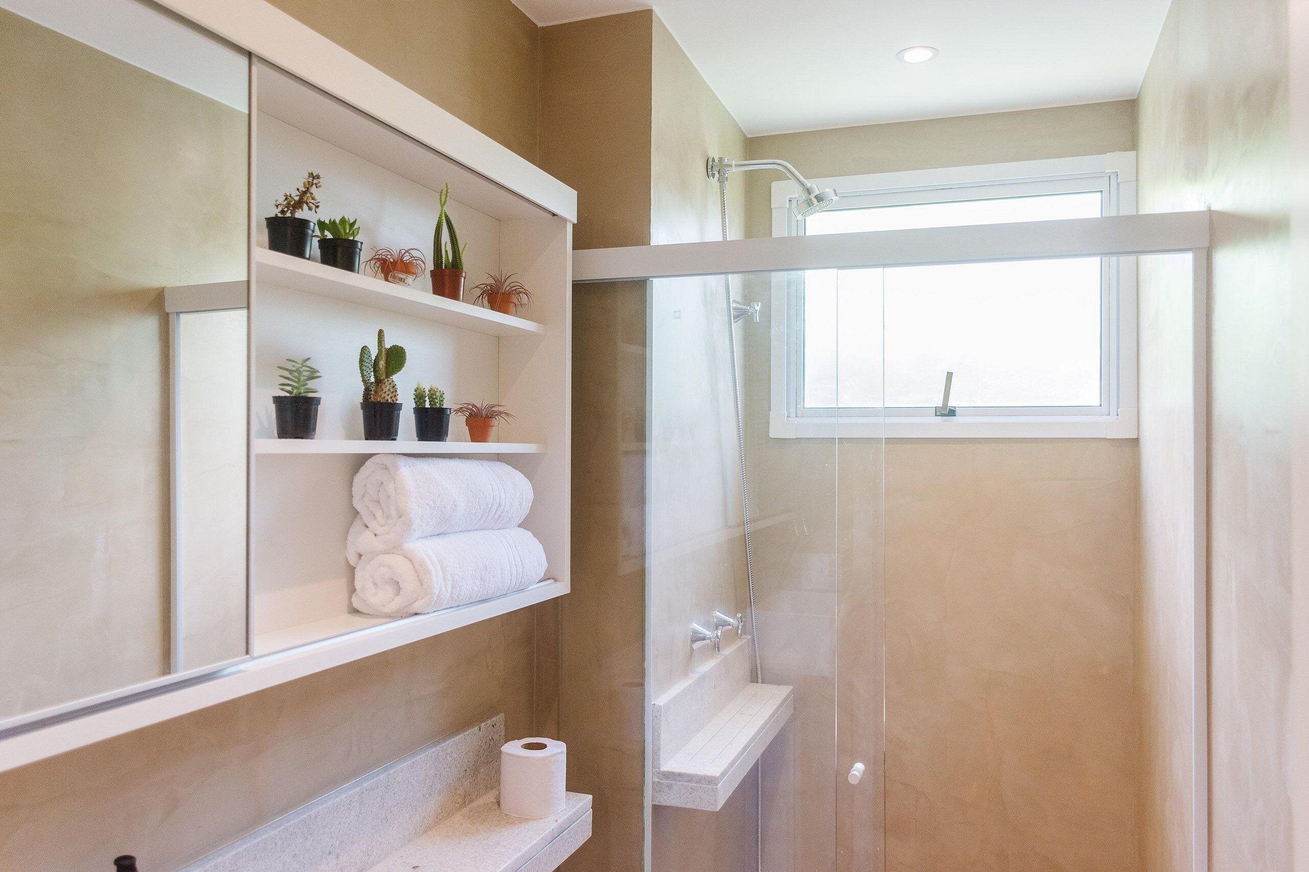 A bathroom with a glass-enclosed shower, a window, shelves with plants and towels, and a toilet paper roll.