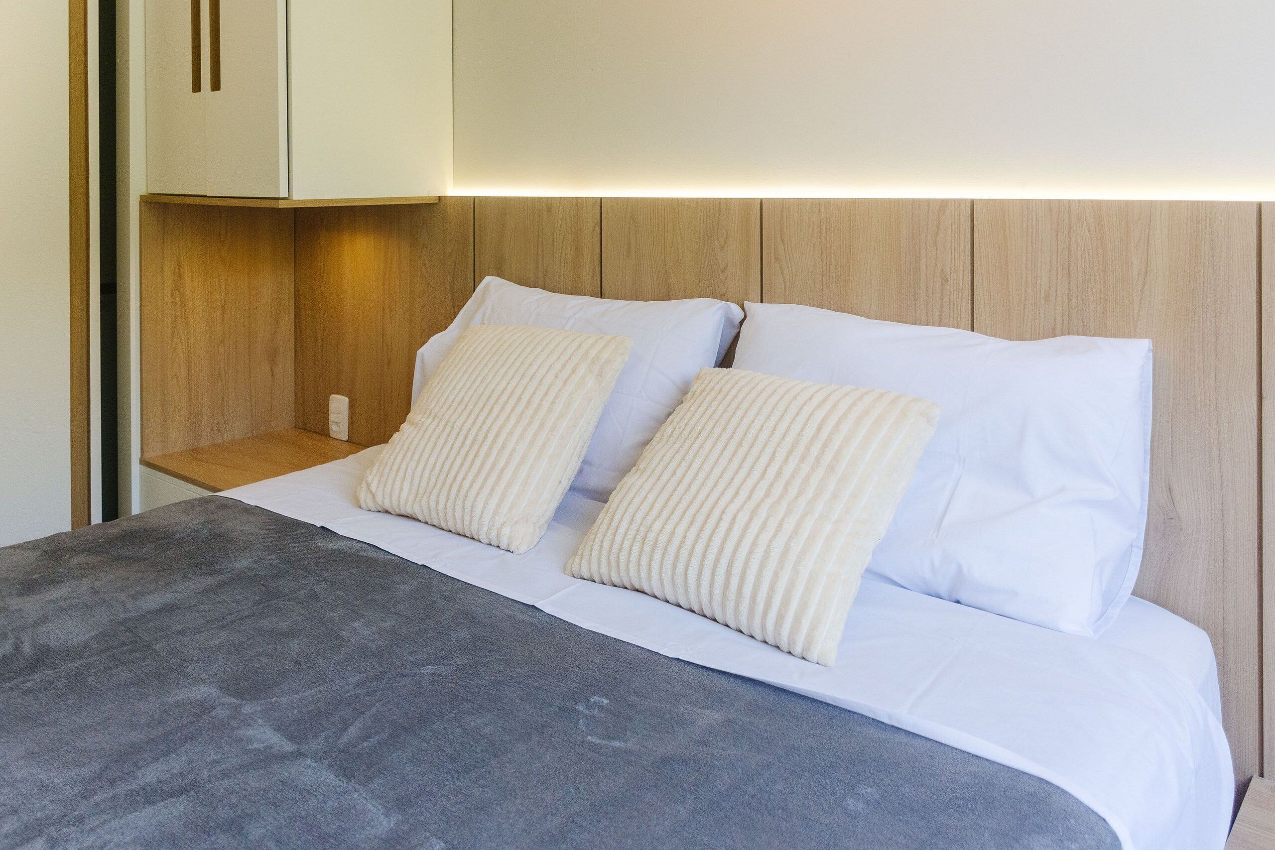 A bed with white linens and two pillows, a wooden headboard, and a bedside table with a lamp.
