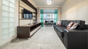 Ein modernes Wohnzimmer mit einer schwarzen Ledersofa, einem flachbildfernseher an einem Holzkasten, einem Essbereich mit Tisch und Stühlen und einem großen Fenster mit Milchglas.