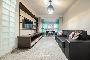 Una sala moderna con un sofá de cuero negro, un televisor de pantalla plana montado en un mueble de madera, un comedor con mesa y sillas, y un ventanal con paneles de vidrio esmerilado.