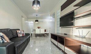Una sala de estar moderna con un sofá de cuero negro, una mesa de comedor con sillas, un televisor montado en un mueble de madera y una araña.