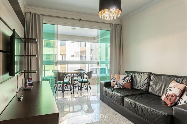 A modern living room with a leather sofa, a flat-screen TV, a dining area with a table and chairs, and large windows offering a view of the cityscape.