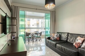 A modern living room with a leather sofa, a flat-screen TV, a dining area with a table and chairs, and large windows offering a view of the cityscape. - Apt W/balcony 160m Bombas Beach Col102 (Bombinhas)