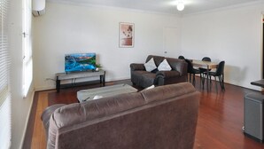 Uma sala de estar com um sofá de couro marrom, uma TV em um suporte de madeira, uma mesa de jantar com cadeiras e uma porta ao fundo.