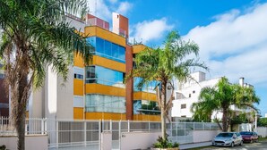 A modern building with a white and yellow facade, large windows, and a gated driveway with parked cars.