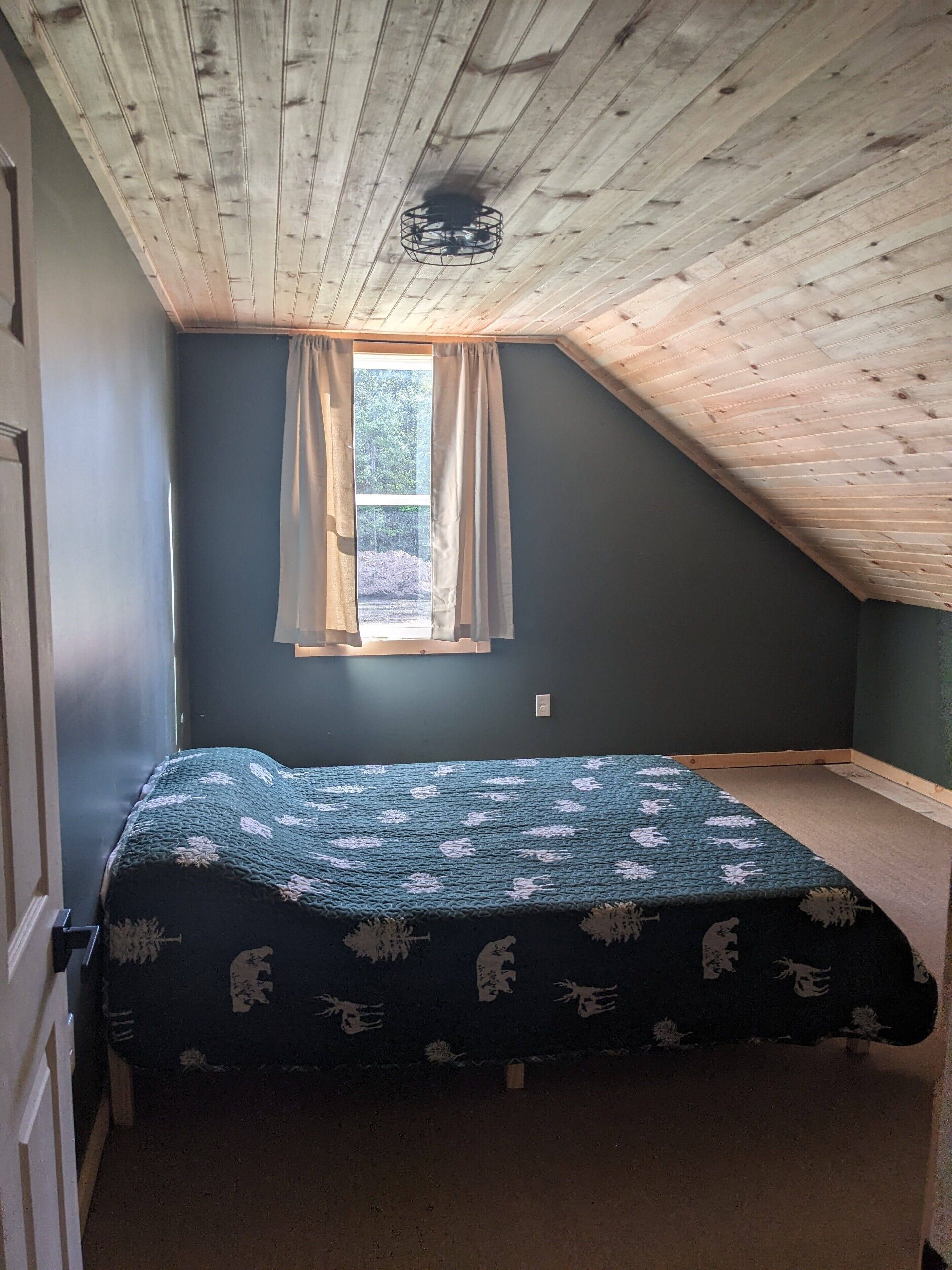 Un dormitorio con techo inclinado, una ventana con cortinas, una cama con colcha estampada y un ventilador de techo.