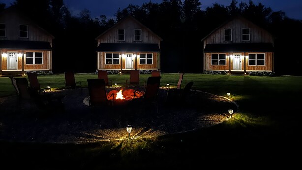 Three wooden cabins with a central fire pit, surrounded by chairs and a well-lit lawn. - The Cabins at Timber Ridge - #3 - Brand new cabins nestled in the woods of NH (Tamworth)