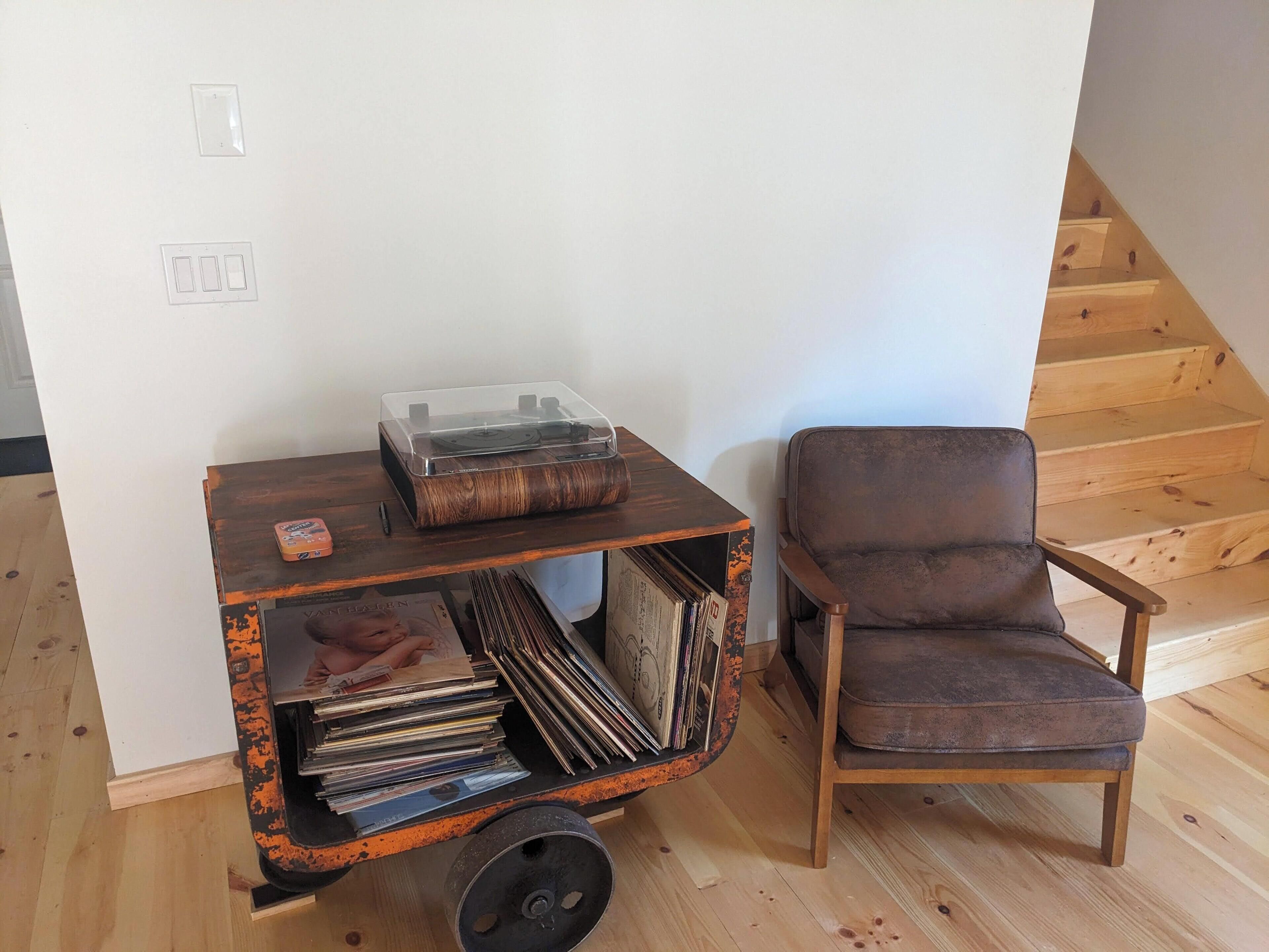 A wooden chair next to a small cart with a record player and vinyl records.