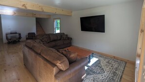 A spacious living room with a sectional sofa, a flat-screen TV mounted on the wall, and a wooden floor. - The Cabins at Timber Ridge - #3 - Brand new cabins nestled in the woods of NH (Tamworth)