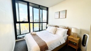 Una habitación de hotel moderna con un ventanal que ofrece vistas a la ciudad, una cama bien hecha con ropa blanca y una mesita de noche con una lámpara.
