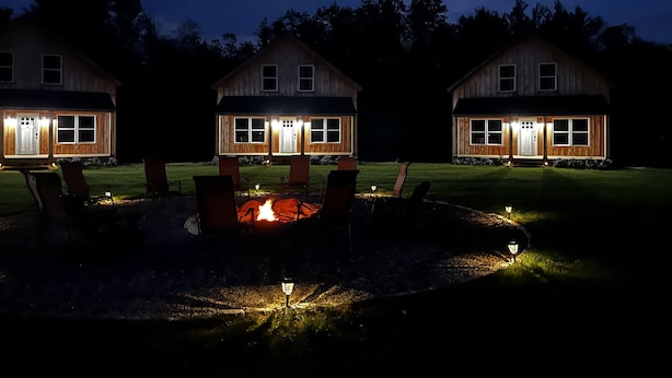 Tres cabañas de madera con un hogar central, rodeadas de sillas y un césped bien iluminado.