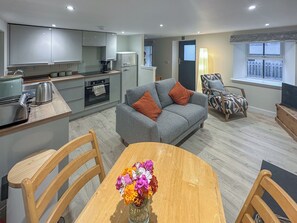 A modern kitchen with a dining area, a grey sofa with orange cushions, and a wooden table with a vase of flowers.