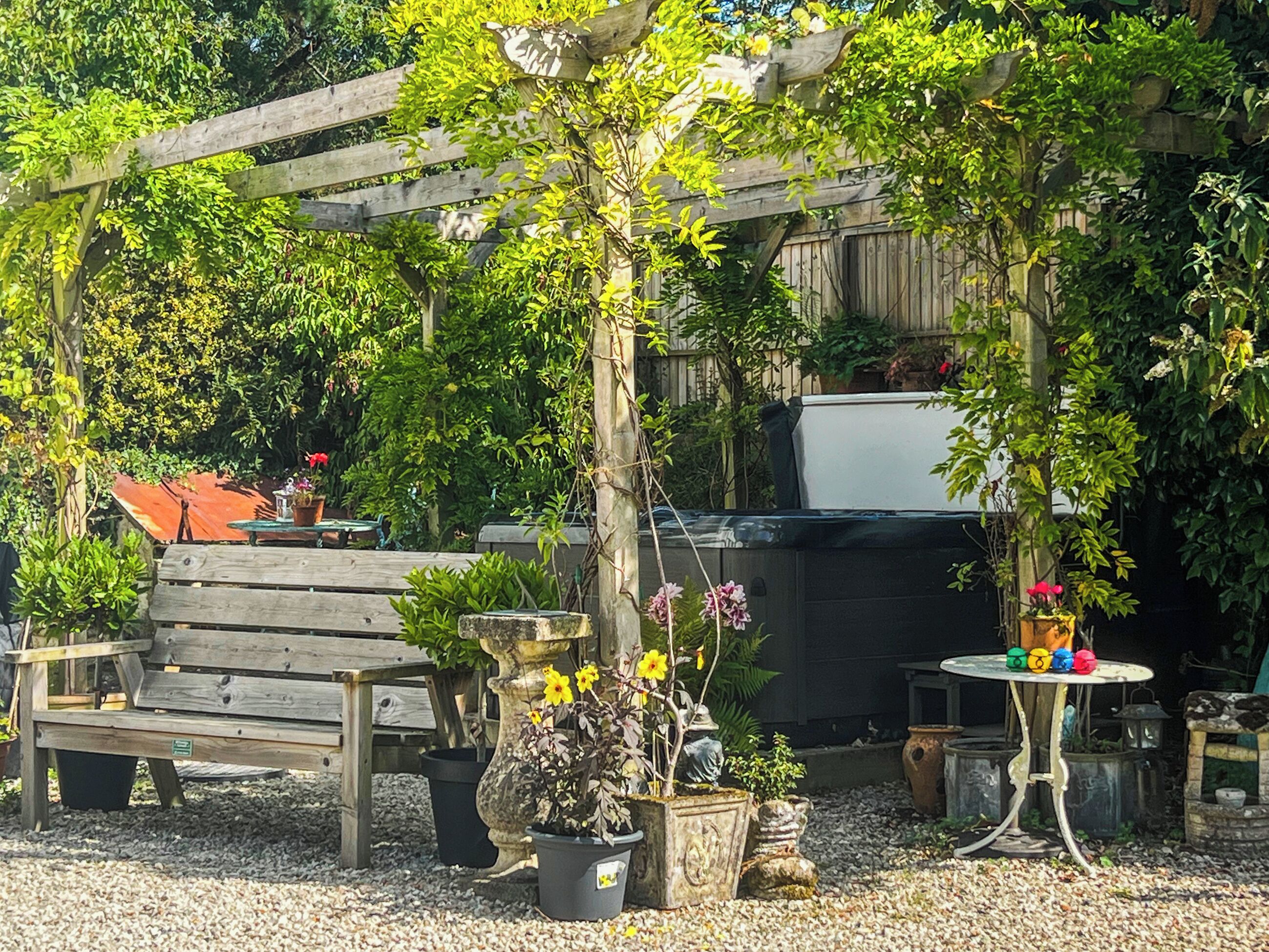 En have med et træpergola, plantekrukker, en træbænk og et rundt bord med farverige genstande.