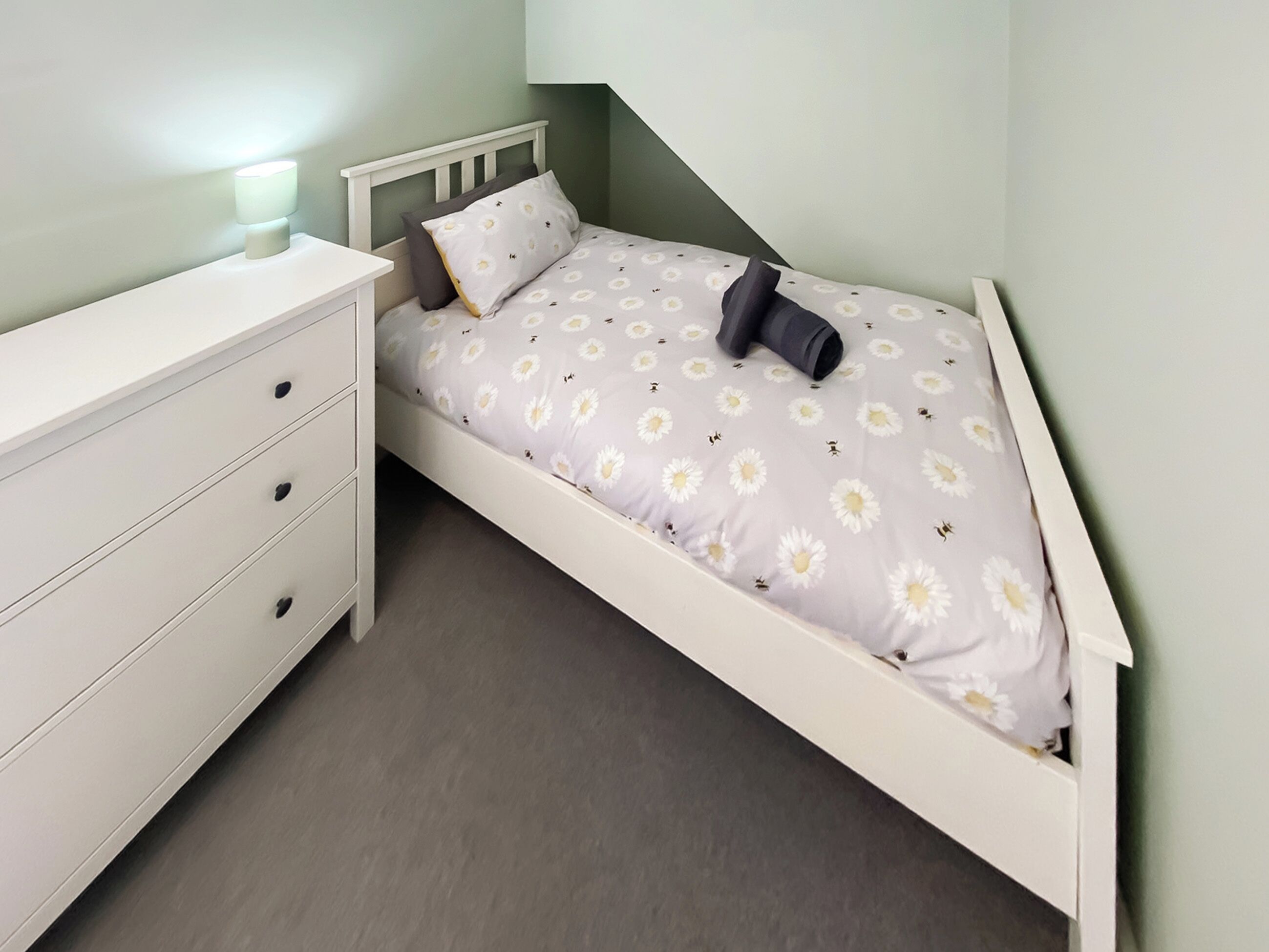 A small, single-bed room with a white bed, a bedside table, and a drawer unit.