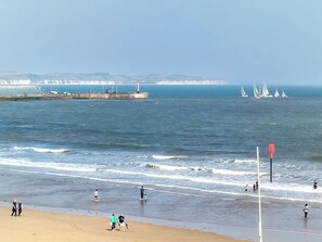 Una playa con gente caminando y parada cerca del agua. Se ven barcos a lo lejos y un faro en el horizonte.