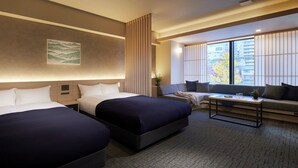 Ett hotellrum med två sängar, en soffa, ett litet bord och ett stort fönster med utsikt över byggnader och träd.