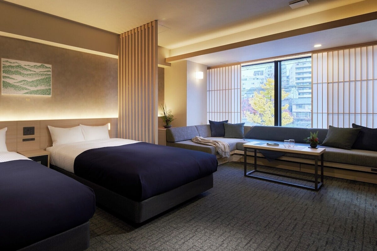 A hotel room with two beds, a sofa, a small table, and a large window with a view of buildings and trees.