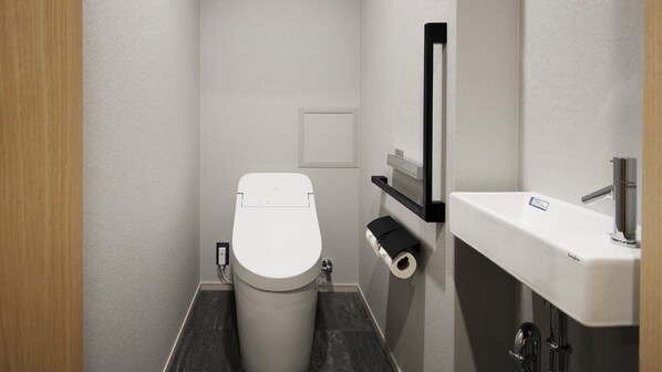 Une petite salle de bain avec des toilettes blanches, un porte-rouleau de papier toilette noir et un lavabo blanc doté d’un robinet chromé.