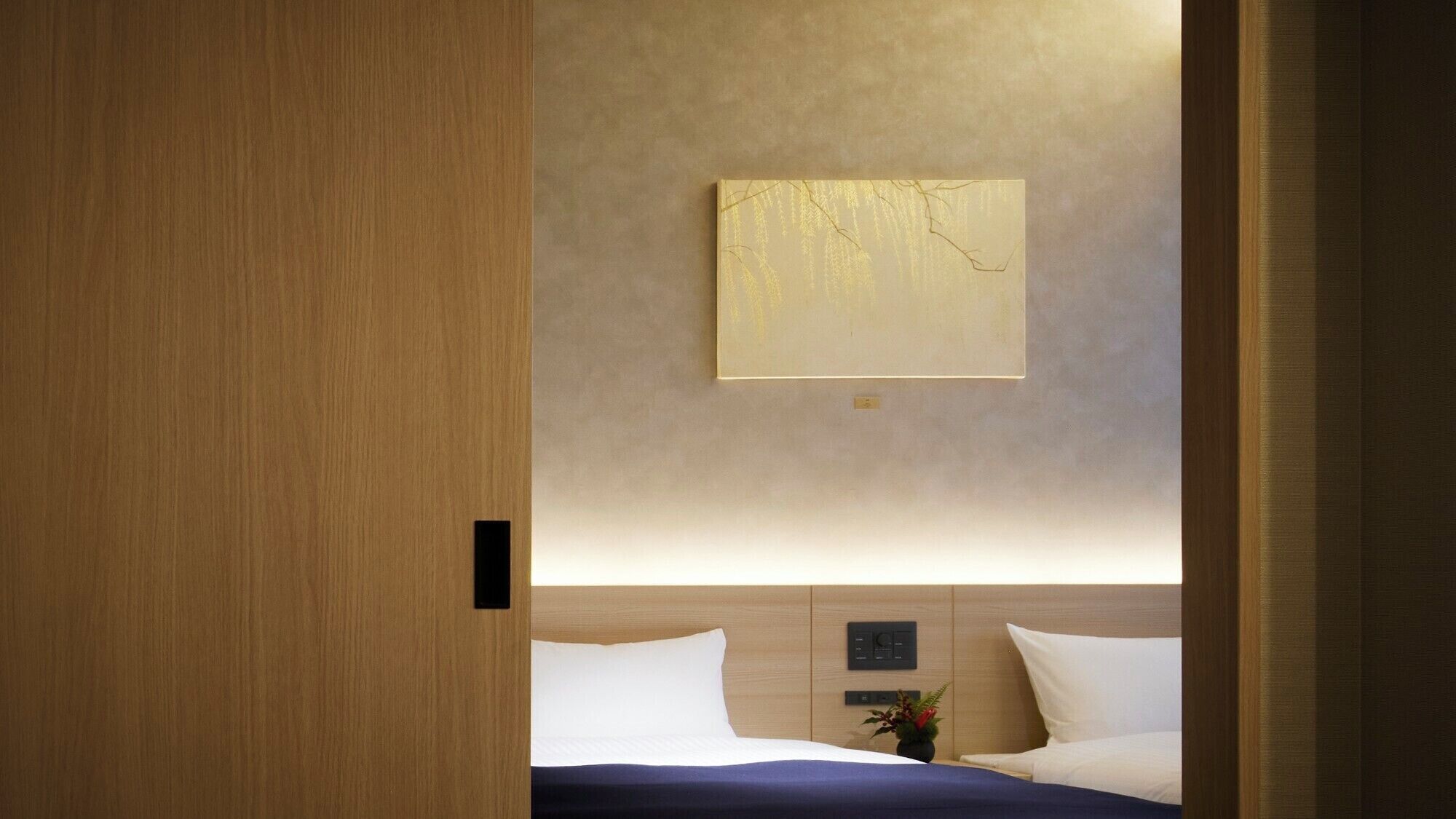 Una habitación de hotel moderna con puerta de madera, cama con ropa blanca, mesita de noche con un florero y una lámpara fijada a la pared.