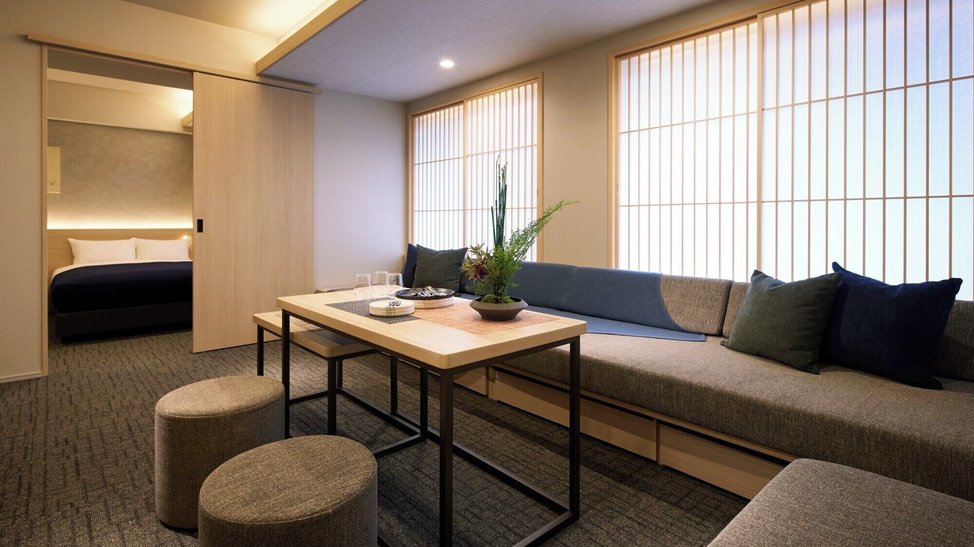 Quarto de hotel compacto com cama, sofá, mesinha com uma planta e janela com persianas verticais.
