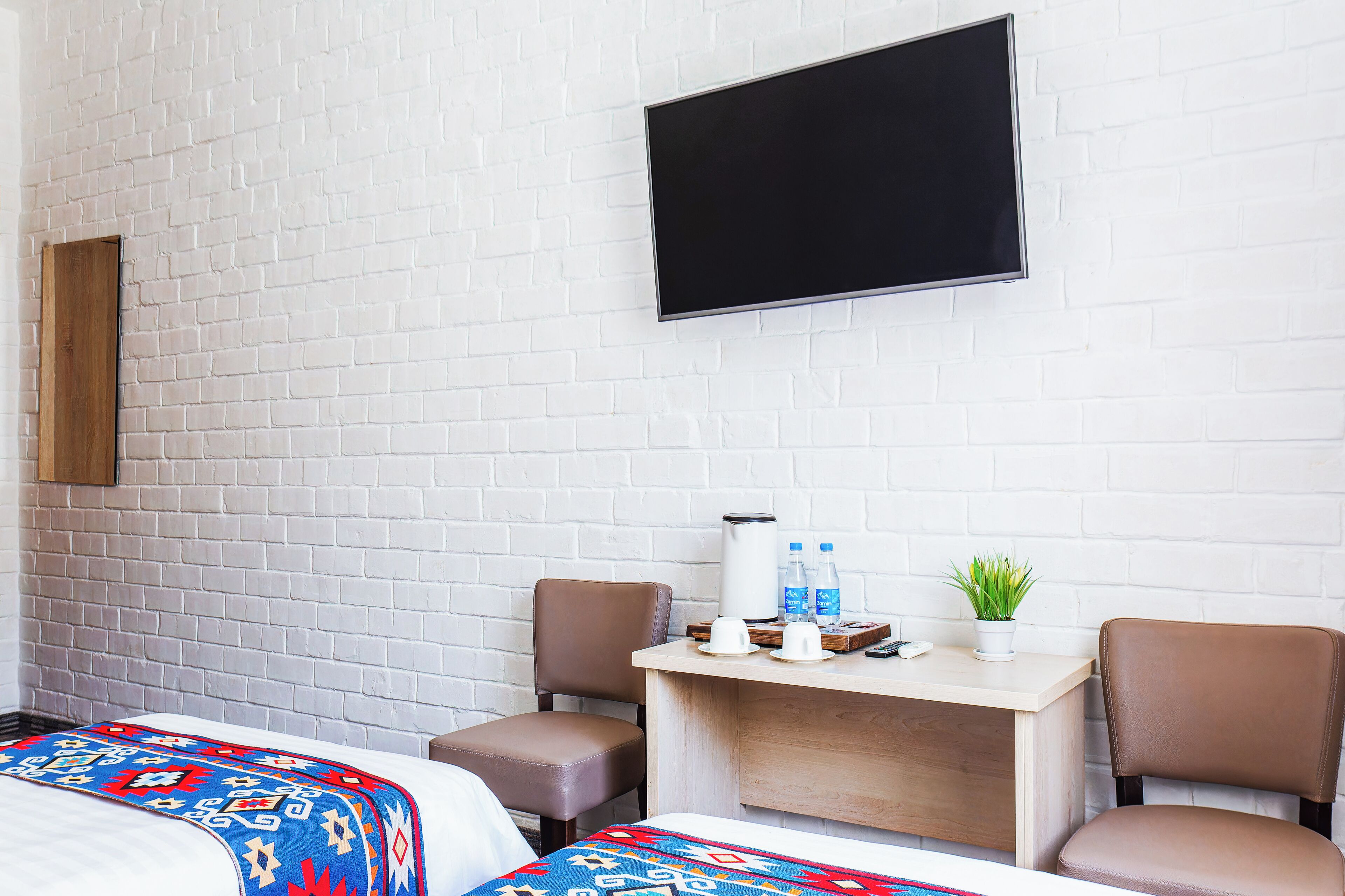 Une chambre d’hôtel avec un mur en briques blanches, un téléviseur à écran plat fixé au-dessus d’un bureau en bois, deux fauteuils en cuir marron et un lit recouvert d’une couverture colorée.
