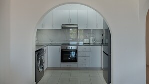 A compact kitchen with a washing machine, oven, and a coffee maker. - Phaedrus Living: Golden Seaview Residence (Paphos)