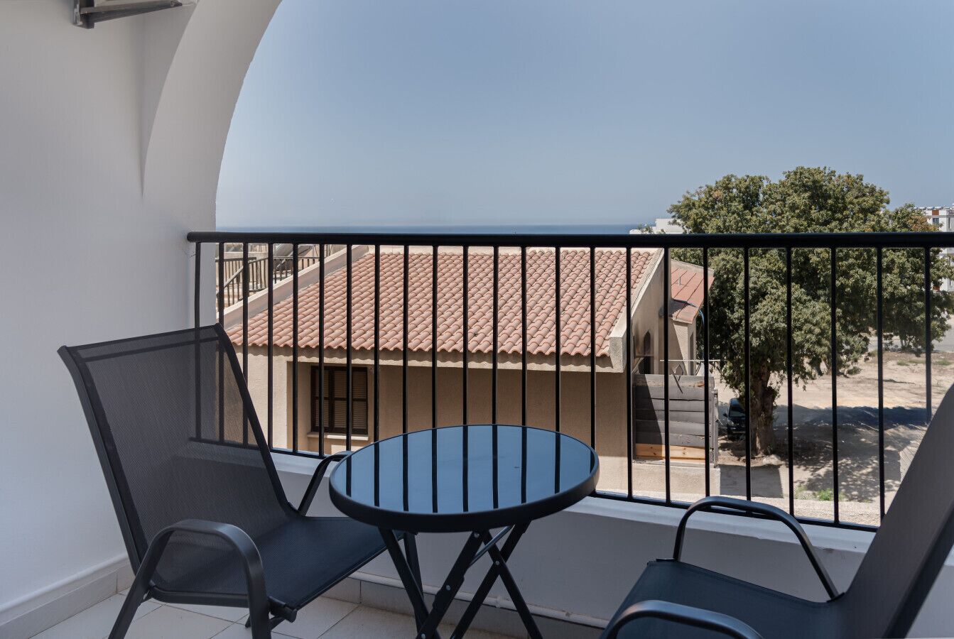 A balcony with a round table and two chairs, a view of a building with a tiled roof, and a tree in the background.