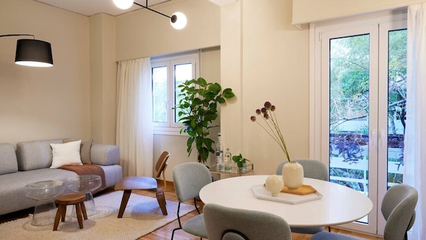 Een moderne woonkamer met een grijze bank, een ronde salontafel en een eettafel met stoelen. Er is een groot raam met gordijnen, een potplant en een vaas met bloemen op de eettafel.
