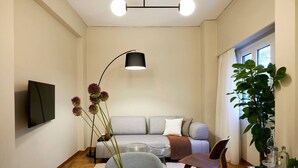 Een moderne woonkamer met een grijze bank, een ronde salontafel, een aan de muur bevestigde televisie, een vloerlamp en een grote potplant.