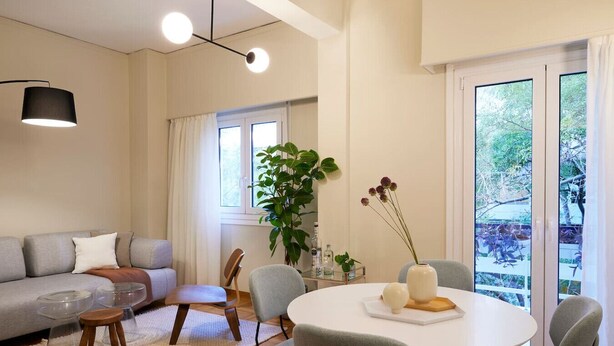 Una sala de estar moderna con un sofá gris, una mesa de comedor redonda y un ventanal con vistas al verdor.