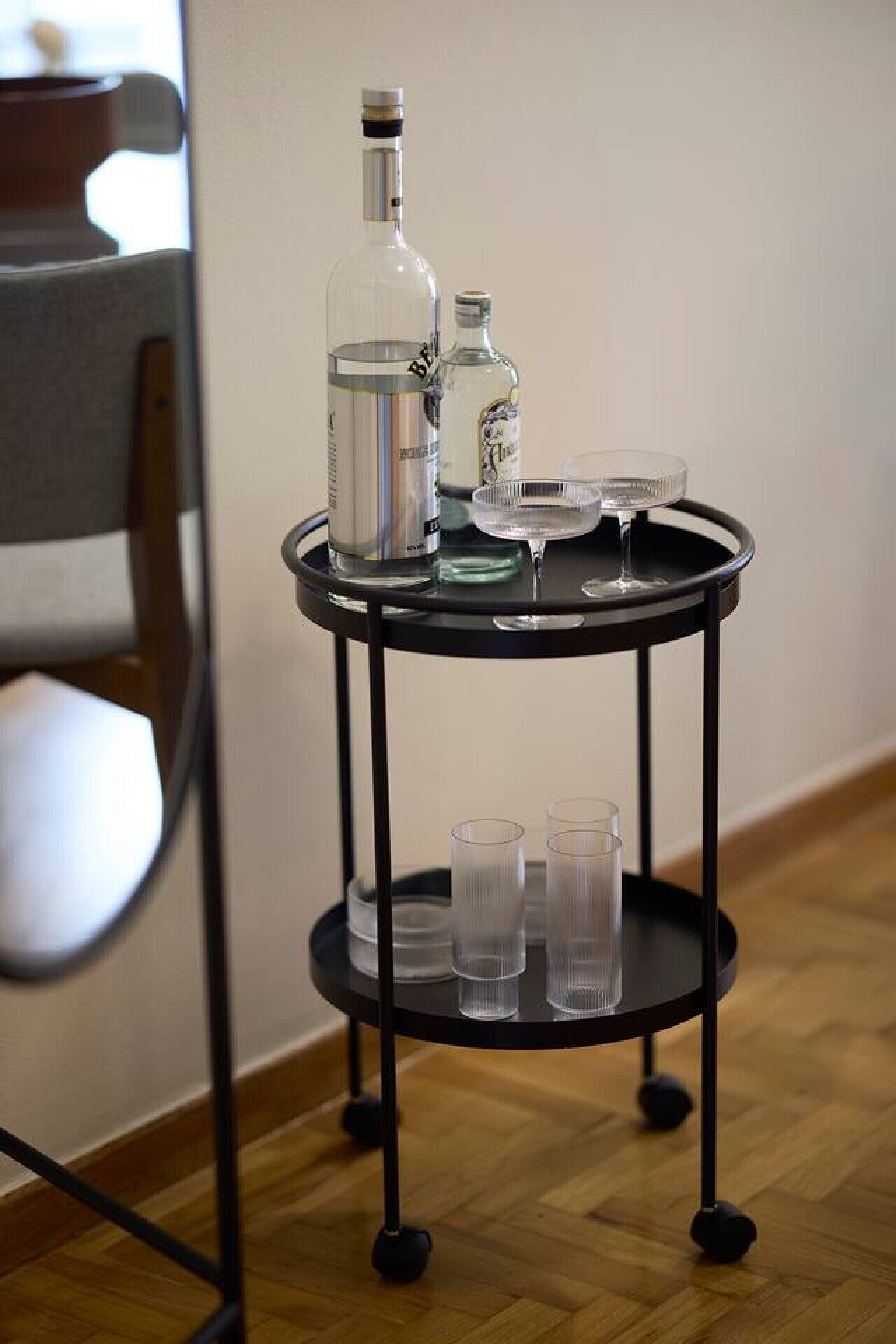 Un carro metálico negro con una botella, un vaso y varios vasos exprés sobre un piso de madera.