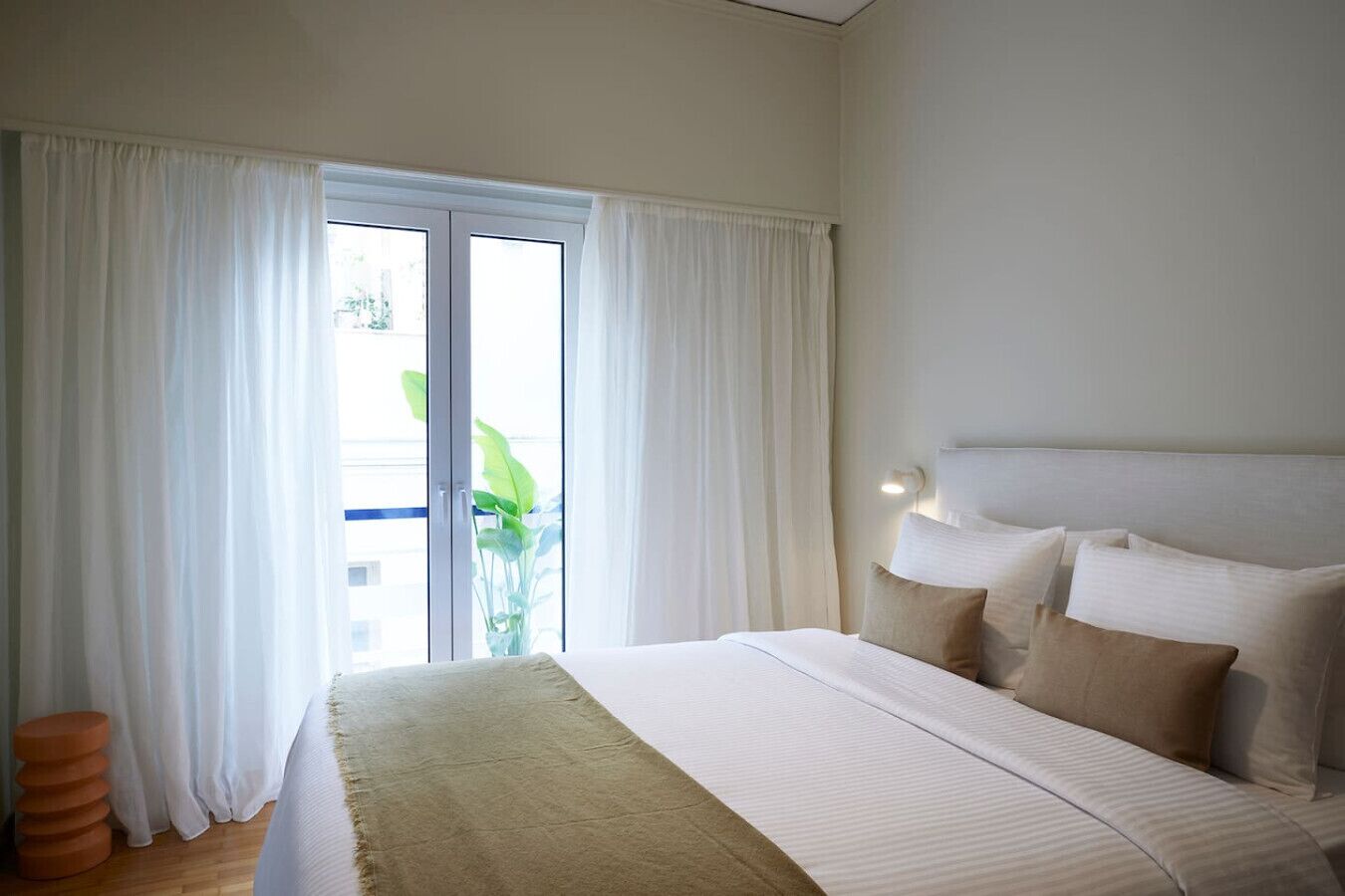 A hotel room with a large bed, white curtains, a potted plant, and a view of the outdoors.