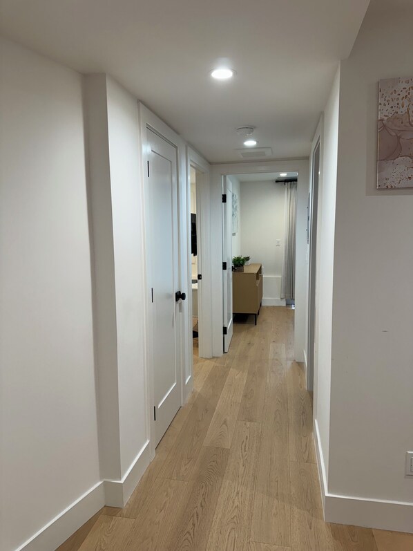 Un couloir aux murs blancs et au plancher en bois, qui mène à une pièce contenant un lit et une fenêtre.