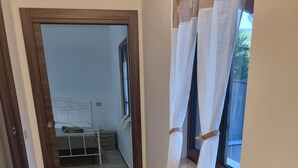 Un couloir au plancher de bois menant à une chambre à coucher équipée d’un lit et d’une fenêtre avec des rideaux.