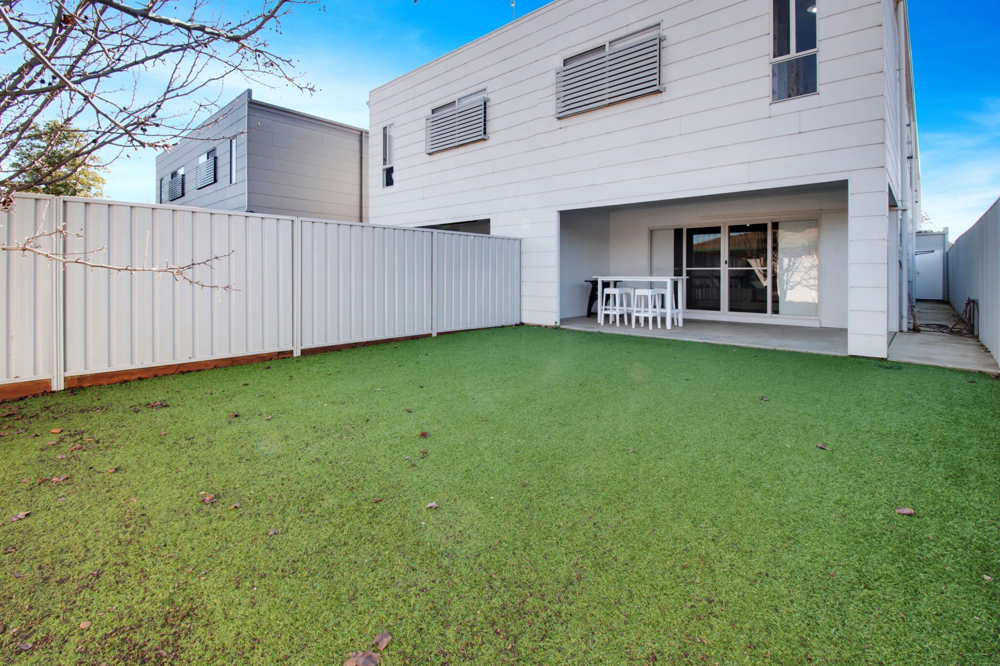 A modern two-story house with a white exterior, a fenced backyard, and a patio area with outdoor furniture.