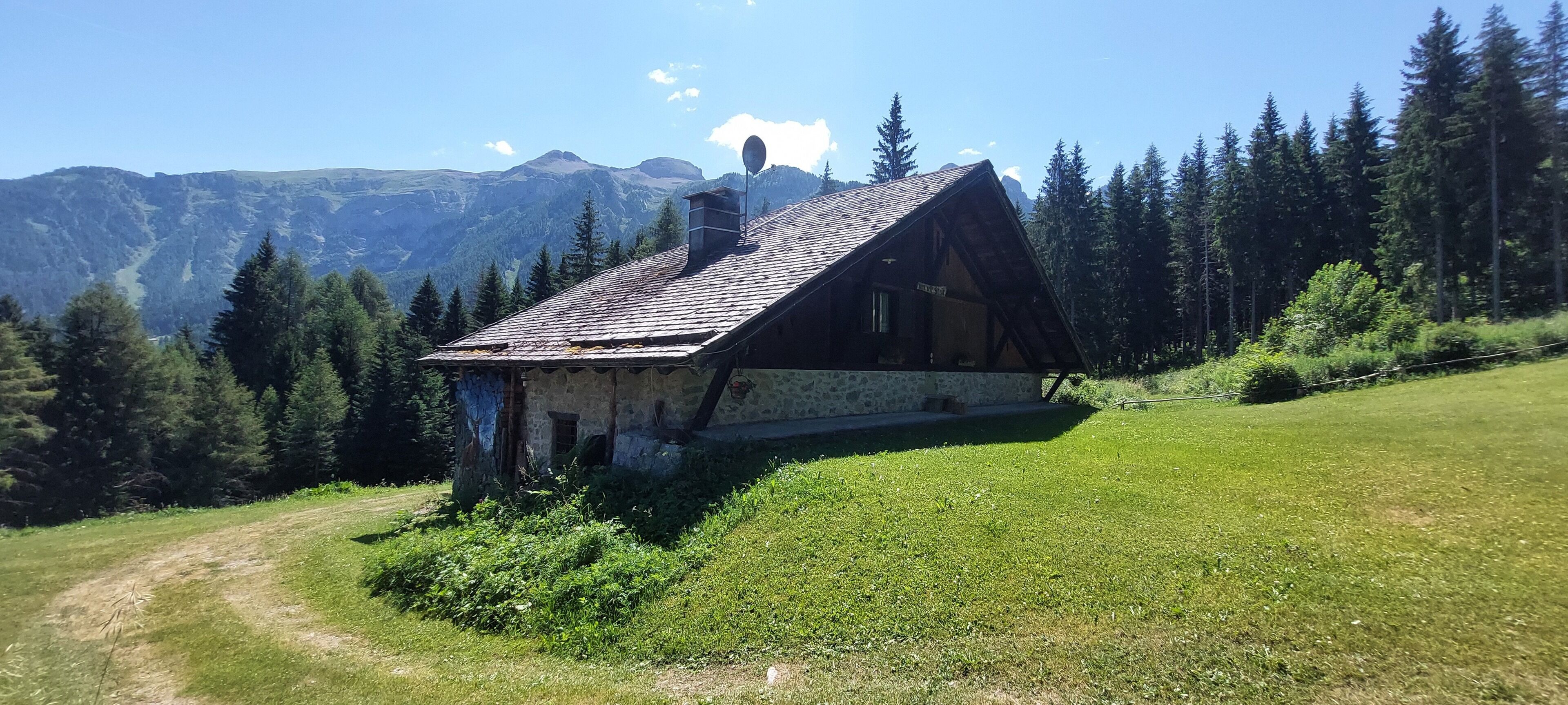 A rustic mountain cabin with a stone foundation and wooden beams, surrounded by lush greenery and mountains in the background.