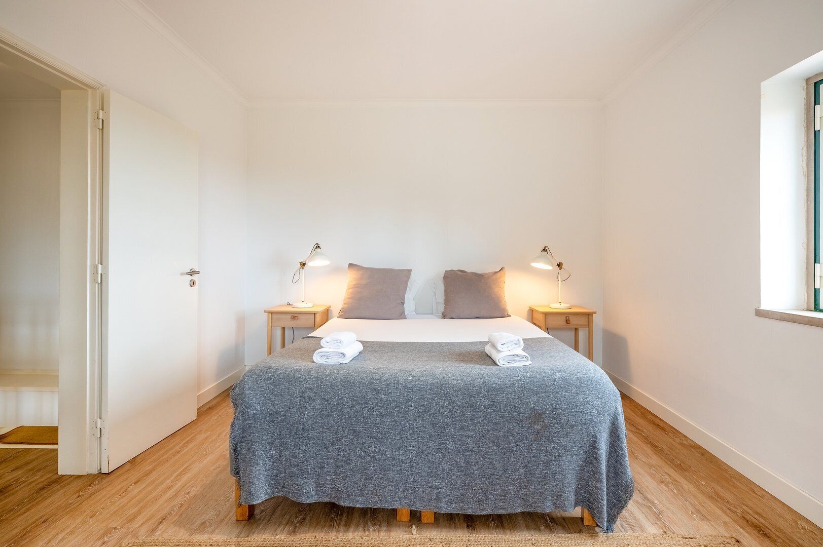Une petite chambre à coucher, bien rangée, avec un lit, deux tables de chevet dotées de lampes, une fenêtre et une porte.