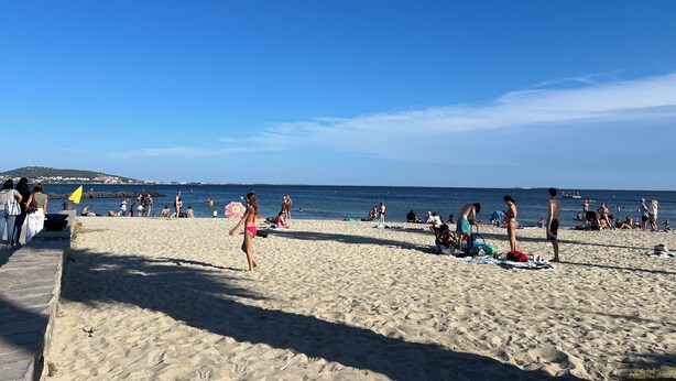 Uma praia com pessoas se banhando e tomando sol, céu claro e um litoral distante.