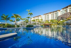Una piscina de resort con palmeras, sillas de descanso y un edificio grande al fondo.